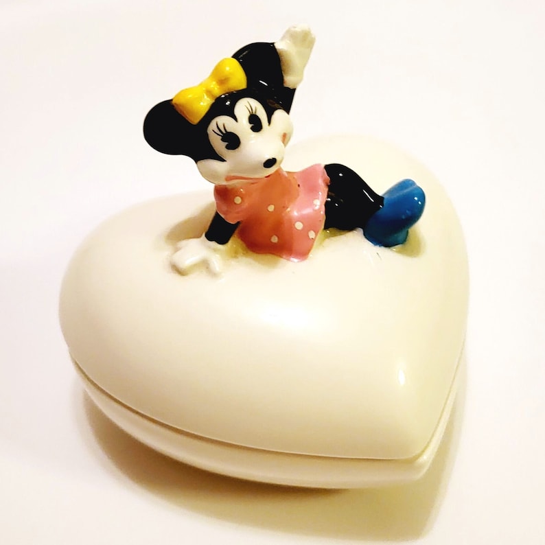 May include: A white heart-shaped ceramic trinket box with a Minnie Mouse figurine on top. Minnie is wearing a pink polka dot dress, yellow bow, and blue shoes. She is waving with her right hand.