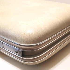 May include: Cream-colored vintage suitcase with a textured surface and silver-toned metal trim. The suitcase has a rectangular shape with rounded corners and a secure latch. A classic travel accessory.