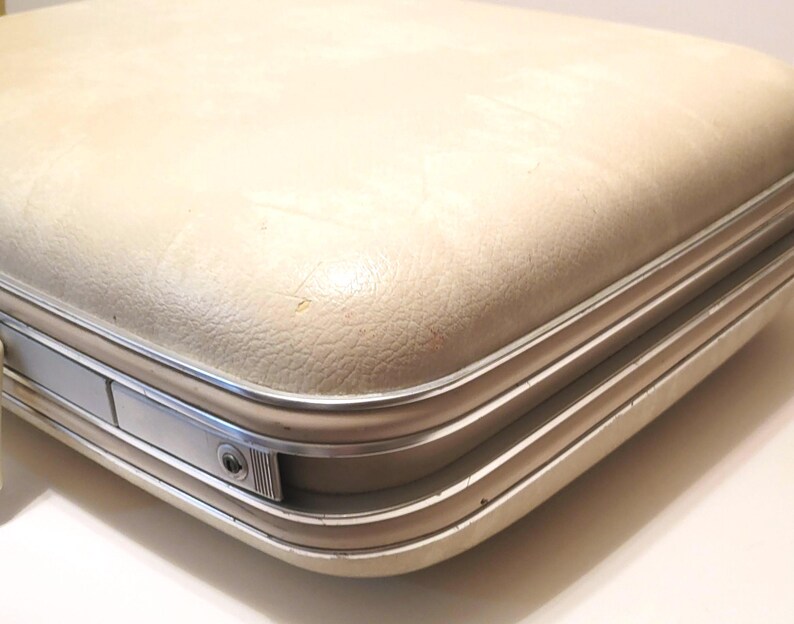 May include: Cream-colored vintage suitcase with a textured surface and silver-toned metal trim. The suitcase has a rectangular shape with rounded corners and a secure latch. A classic travel accessory.