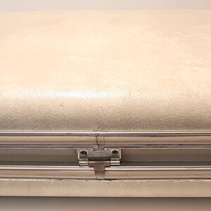 May include: A vintage cream-colored suitcase with rounded edges and a textured surface. The suitcase has a silver-colored metal frame and hardware, including hinges and latches. Small, rounded feet are visible.