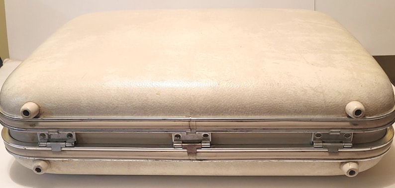 May include: A vintage cream-colored suitcase with rounded edges and a textured surface. The suitcase has a silver-colored metal frame and hardware, including hinges and latches. Small, rounded feet are visible.