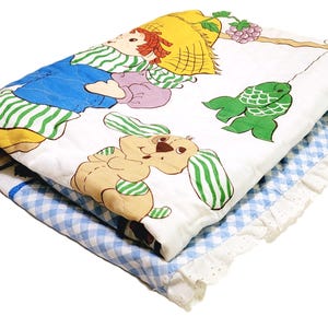May include: A white quilted blanket with a blue and green gingham border. The blanket features a cartoon illustration of a girl with a straw hat, a dog, and a fish.