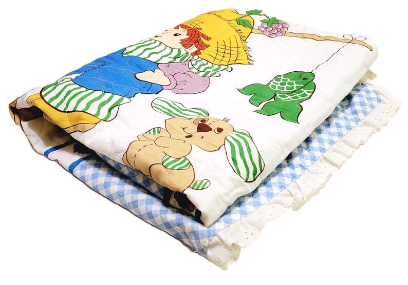May include: A white quilted blanket with a blue and green gingham border. The blanket features a cartoon illustration of a girl with a straw hat, a dog, and a fish.