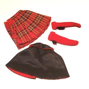 May include: A doll-sized outfit featuring a red plaid skirt, a black cape with a red trim, and two red leg warmers. The skirt has a pleated design with yellow and purple accents. The cape is lined with red fabric.