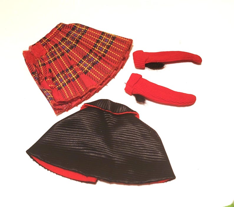 May include: A doll-sized outfit featuring a red plaid skirt, a black cape with a red trim, and two red leg warmers. The skirt has a pleated design with yellow and purple accents. The cape is lined with red fabric.