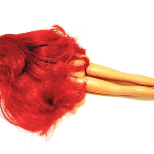 May include: A red-haired doll lying on its back with its legs extended. The doll has a tan complexion and is wearing no clothing.