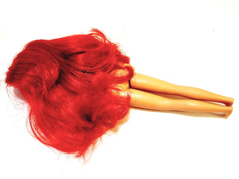 May include: A red-haired doll lying on its back with its legs extended. The doll has a tan complexion and is wearing no clothing.