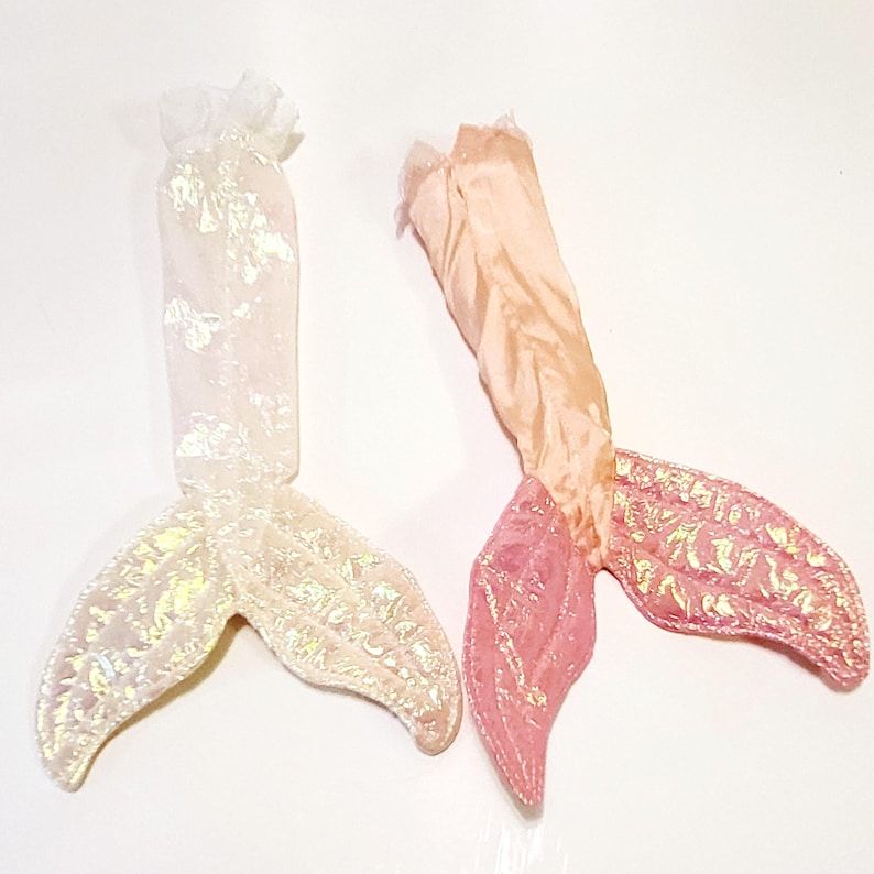 May include: Two mermaid tails made of fabric. One tail is white with iridescent glitter and the other is pink with gold glitter.