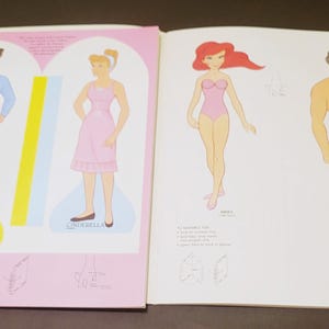 Pode incluir: Livro de bonecas de papel vintage com ilustra&ccedil;&otilde;es de personagens de desenhos animados. O livro mostra o Pr&iacute;ncipe Encantado em azul, Cinderela em rosa, Ariel em um mai&ocirc; rosa e um homem de shorts vermelhos.