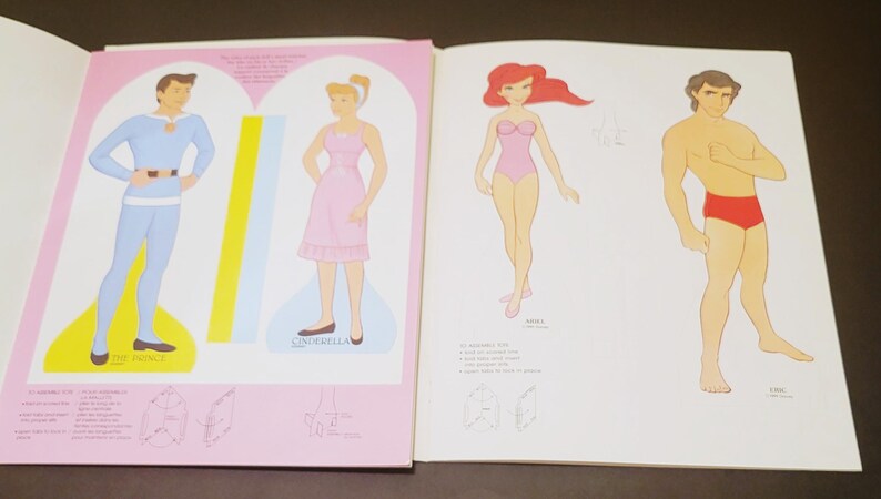 Pode incluir: Livro de bonecas de papel vintage com ilustra&ccedil;&otilde;es de personagens de desenhos animados. O livro mostra o Pr&iacute;ncipe Encantado em azul, Cinderela em rosa, Ariel em um mai&ocirc; rosa e um homem de shorts vermelhos.