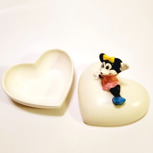 May include: A white ceramic heart-shaped trinket box with a lid. The box features a small, hand-painted figurine of a cartoon mouse with black ears, a yellow bow, a pink dress with white polka dots, and blue shoes.