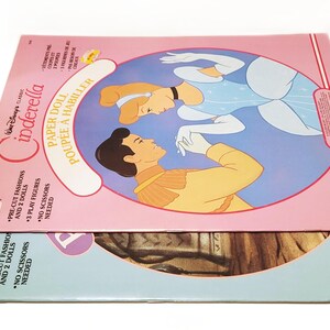 Pode incluir: Conjunto de bonecas de papel Cinderela da Disney vintage. A caixa rosa apresenta Cinderela e o Pr&iacute;ncipe Encantado, com o texto "PAPER DOLL POUPEE A HABILLER". O conjunto inclui modas pr&eacute;-cortadas, 2 bonecas e 3 figuras de jogo. A caixa inferior &eacute; azul e castanha.
