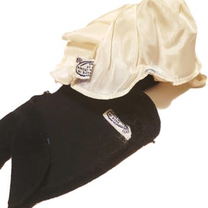 May include: A black velvet hat with a cream-colored satin lining. The hat has a rounded crown and a brim that curves upwards. A small tag is sewn on the hat with text that reads "MADE IN CROWN".