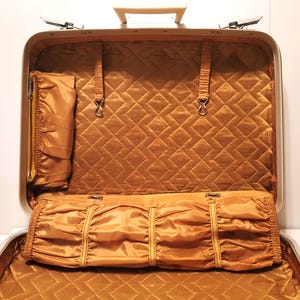 May include: Open vintage suitcase with a gold interior. The interior features a quilted pattern and several compartments, including a zippered pocket and a pleated section. The suitcase has a silver frame and a cream-colored handle.