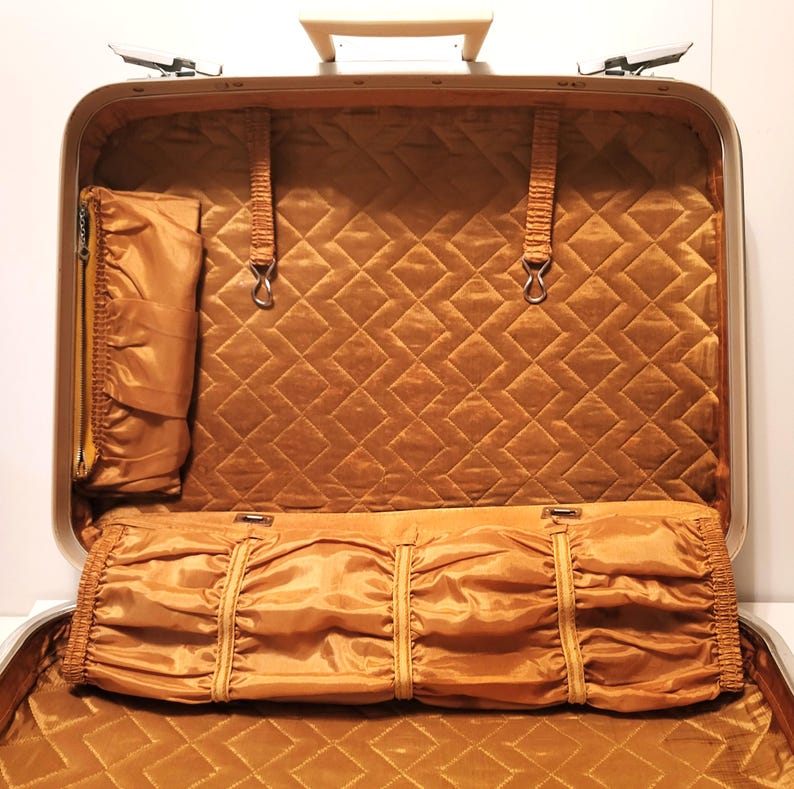 May include: Open vintage suitcase with a gold interior. The interior features a quilted pattern and several compartments, including a zippered pocket and a pleated section. The suitcase has a silver frame and a cream-colored handle.