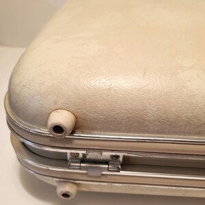 May include: Close-up of a vintage off-white suitcase with rounded edges. The suitcase has a silver metal frame and hinges, with two small, white, cylindrical feet and a handle. The surface shows signs of wear.