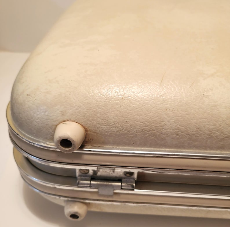 May include: Close-up of a vintage off-white suitcase with rounded edges. The suitcase has a silver metal frame and hinges, with two small, white, cylindrical feet and a handle. The surface shows signs of wear.