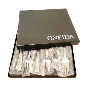Peut inclure: Un ensemble de fourchettes argentées dans une boîte noire. Le couvercle de la boîte est ouvert, révélant les fourchettes emballées individuellement dans du plastique transparent. La marque "ONEIDA" est imprimée en blanc sur le couvercle de la boîte. Les fourchettes sont rangées à l'intérieur.