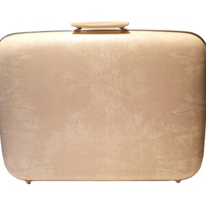 May include: A vintage, rectangular suitcase in a light beige color. The suitcase has a textured surface, rounded corners, and a small, light-colored handle on top. The suitcase has small feet on the bottom.