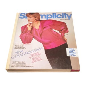 May include: A Simplicity sewing pattern catalog for winter featuring a model in a bright pink long-sleeved blouse and a black skirt. The cover includes text such as Blouse Impact, Festive Attire, New! Les Tous Nouveaux, and Special Pattern Offer.