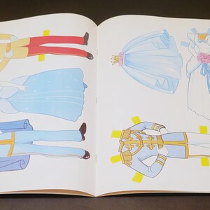 Pode incluir: Livro aberto com ilustra&ccedil;&otilde;es de bonecas de papel. As bonecas incluem um pr&iacute;ncipe com casaco dourado e cal&ccedil;as vermelhas, uma princesa com vestido azul e um pr&iacute;ncipe com uniforme azul. Tamb&eacute;m, um vestido de noiva e um v&eacute;u.