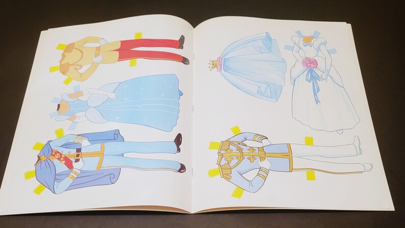 Pode incluir: Livro aberto com ilustra&ccedil;&otilde;es de bonecas de papel. As bonecas incluem um pr&iacute;ncipe com casaco dourado e cal&ccedil;as vermelhas, uma princesa com vestido azul e um pr&iacute;ncipe com uniforme azul. Tamb&eacute;m, um vestido de noiva e um v&eacute;u.