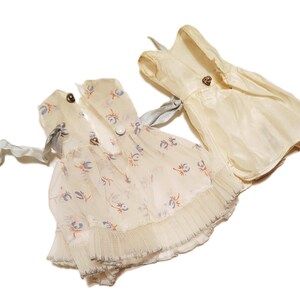 May include: Three vintage doll dresses in cream and off-white. One dress features a floral print with blue and orange accents, while the others are solid. Each dress has decorative buttons and ribbon ties.