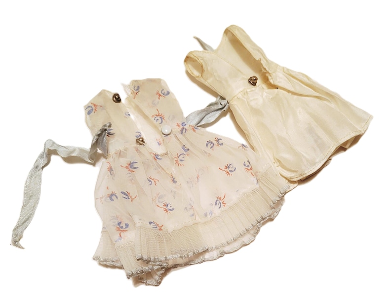 May include: Three vintage doll dresses in cream and off-white. One dress features a floral print with blue and orange accents, while the others are solid. Each dress has decorative buttons and ribbon ties.
