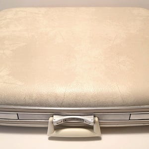 May include: Cream-colored vintage suitcase with a textured surface and silver-toned metal trim. The suitcase features a white handle and two silver latches. A classic travel accessory.