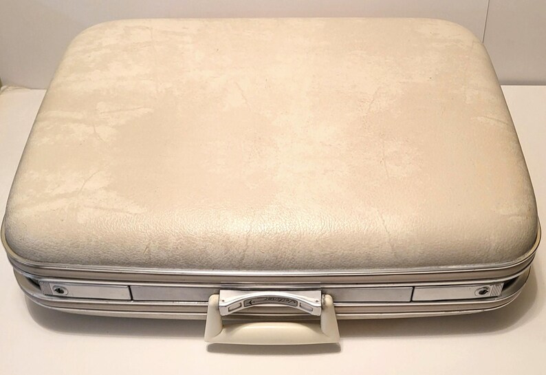 May include: Cream-colored vintage suitcase with a textured surface and silver-toned metal trim. The suitcase features a white handle and two silver latches. A classic travel accessory.