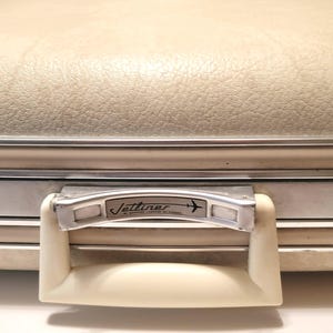 May include: Cream-colored vintage suitcase with a textured surface and silver trim. The handle is off-white with a silver metal plate that reads "Jettliner" and features an airplane graphic. A classic travel accessory.