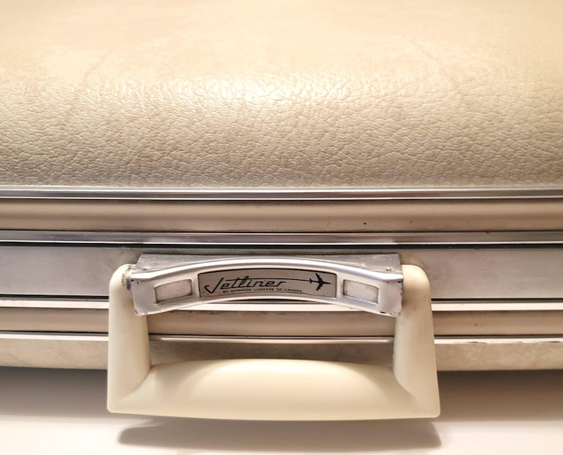 May include: Cream-colored vintage suitcase with a textured surface and silver trim. The handle is off-white with a silver metal plate that reads "Jettliner" and features an airplane graphic. A classic travel accessory.