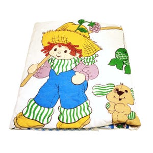 May include: A white quilted blanket with a colorful illustration of a girl with red hair wearing a straw hat and overalls, holding a fishing pole with a fish on the line. A brown and white dog is also pictured on the blanket.