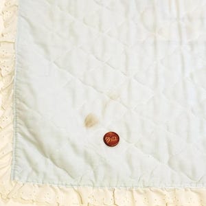 May include: A light blue baby blanket with a white lace border. The blanket has a diamond quilted pattern and a small brown stain.