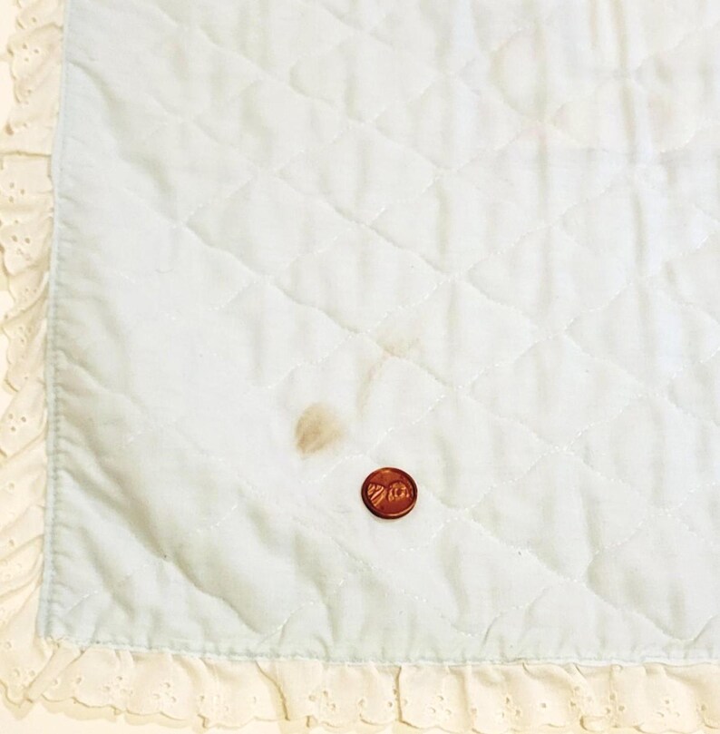 May include: A light blue baby blanket with a white lace border. The blanket has a diamond quilted pattern and a small brown stain.
