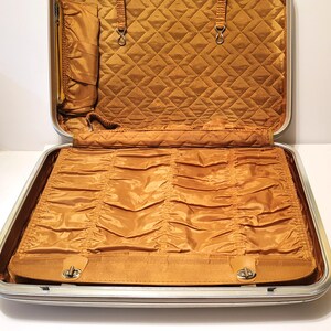 May include: Open vintage suitcase with a silver frame and a mustard-colored interior. The interior features a quilted top section and a bottom section with gathered fabric. The suitcase has a leather strap with a metal clasp.