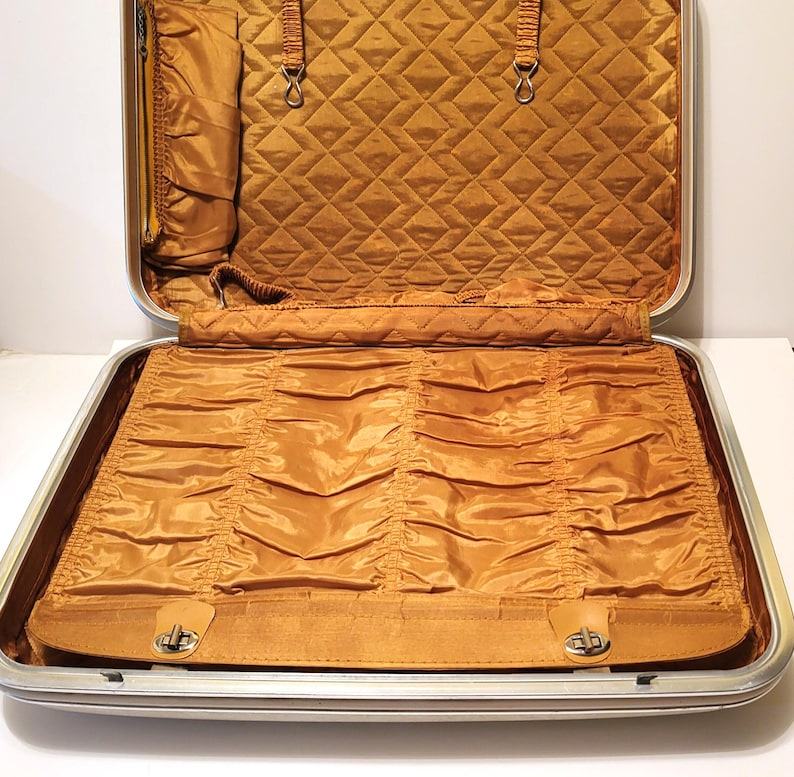 May include: Open vintage suitcase with a silver frame and a mustard-colored interior. The interior features a quilted top section and a bottom section with gathered fabric. The suitcase has a leather strap with a metal clasp.
