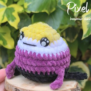 May include: A crocheted plush toy in the shape of a round creature. It features a yellow top, white middle, purple body, and black base. The toy has large eyes and small legs. The text "Pixel handmade plush" is visible in the background.