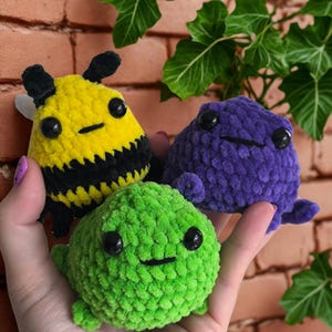 May include: Three crocheted plush toys: a yellow and black bee, a purple blob, and a green blob. Each has large black eyes and a stitched smile. The bee has small wings and antennae. The toys are held in a hand.