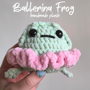 Può includere: Una rana di peluche fatta a mano in verde chiaro con occhi neri e un tutù rosa. Il testo "Ballerina Frog handmade plush" è sopra la rana. La rana è tenuta da una persona.