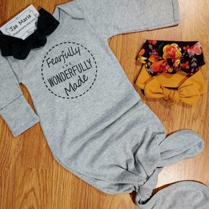 May include: Gray baby gown with long sleeves and a knotted bottom. The gown has a black circle with the text "Fearfully and Wonderfully Made" in white. The image also shows three headbands: one black, one mustard yellow, and one with a floral pattern.