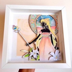 Könnte beinhalten: Gerahmtes Papierkunstwerk mit einer Silhouette einer Figur in einem pfirsichfarbenen Kleid, die einen Zauberstab hält. Der Hintergrund zeigt einen Kreis im Buntglasstil mit Nachthimmel und einem Gebäude. Weiße Lilien sind im Vordergrund.