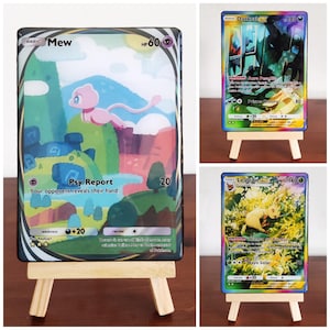 Puede incluir: Tres cartas coleccionables de Pokémon exhibidas en pequeños caballetes de madera. Las cartas presentan ilustraciones coloridas de personajes Pokémon, incluyendo Mew, Darkrai y Leafeon. Las cartas tienen texto con valores de HP y ataque.