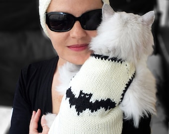 May include: A cream-colored knitted pet sweater featuring a black bat graphic on the back, designed with a ribbed neckline and arm openings. The sweater is crafted from a soft yarn and displays a clear, contrasting bat motif centered on the back panel.