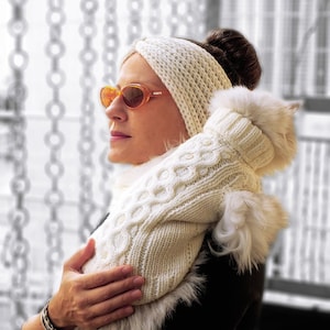 May include: A woman wearing a white knitted headband and sunglasses holds a white knitted sweater with a fur trim.