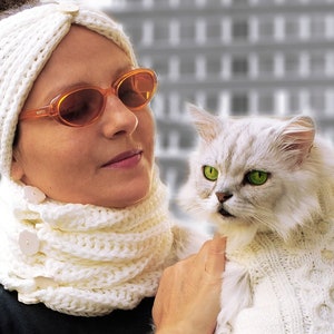 May include: A woman wearing a white knitted headband and scarf is holding a white Persian cat wearing a white knitted sweater. The cat has green eyes and is looking at the camera.