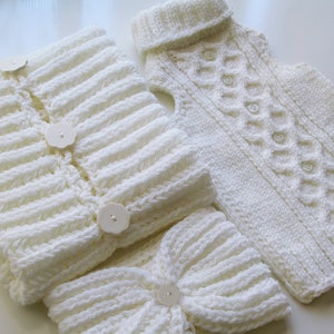 May include: A white knitted baby cardigan, hat, and booties set with button closures. The cardigan has a cable knit pattern and the hat and booties have a ribbed knit pattern.