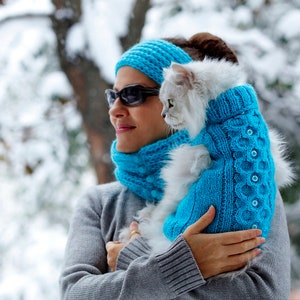 May include: A woman wearing a blue knitted hat and scarf holds a white cat wearing a blue knitted sweater. The cat is looking at the camera.