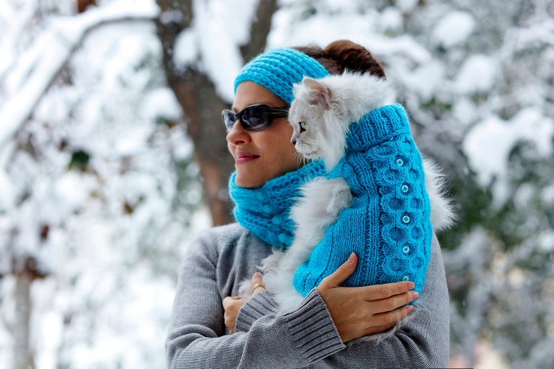 May include: A woman wearing a blue knitted hat and scarf holds a white cat wearing a blue knitted sweater. The cat is looking at the camera.
