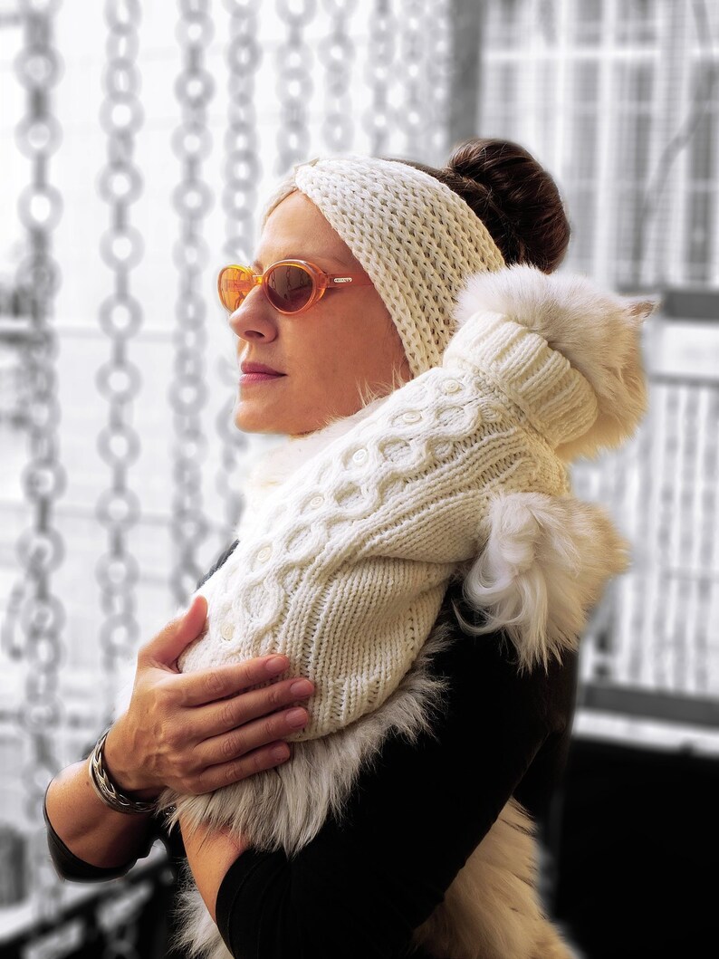 May include: A woman wearing a white knitted headband and sunglasses holds a white knitted sweater with a fur trim.